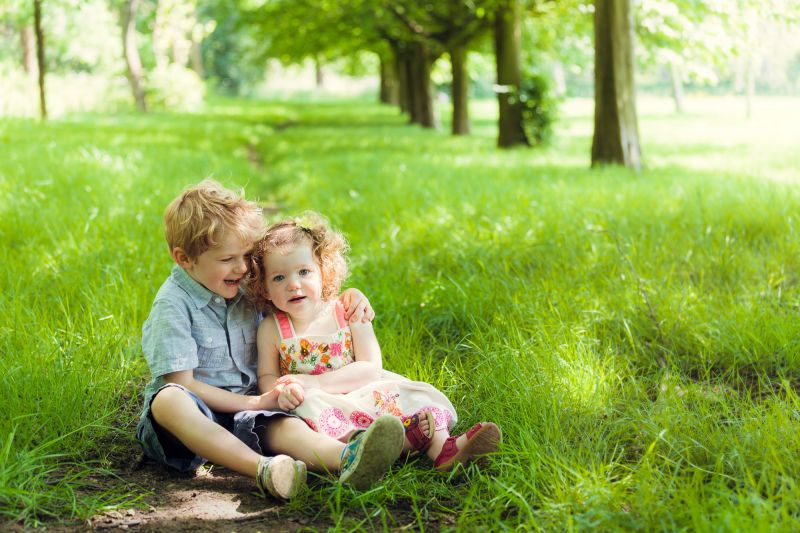 family photography in a park with trees leading off into the background. A brother and sister hold hands sitting down